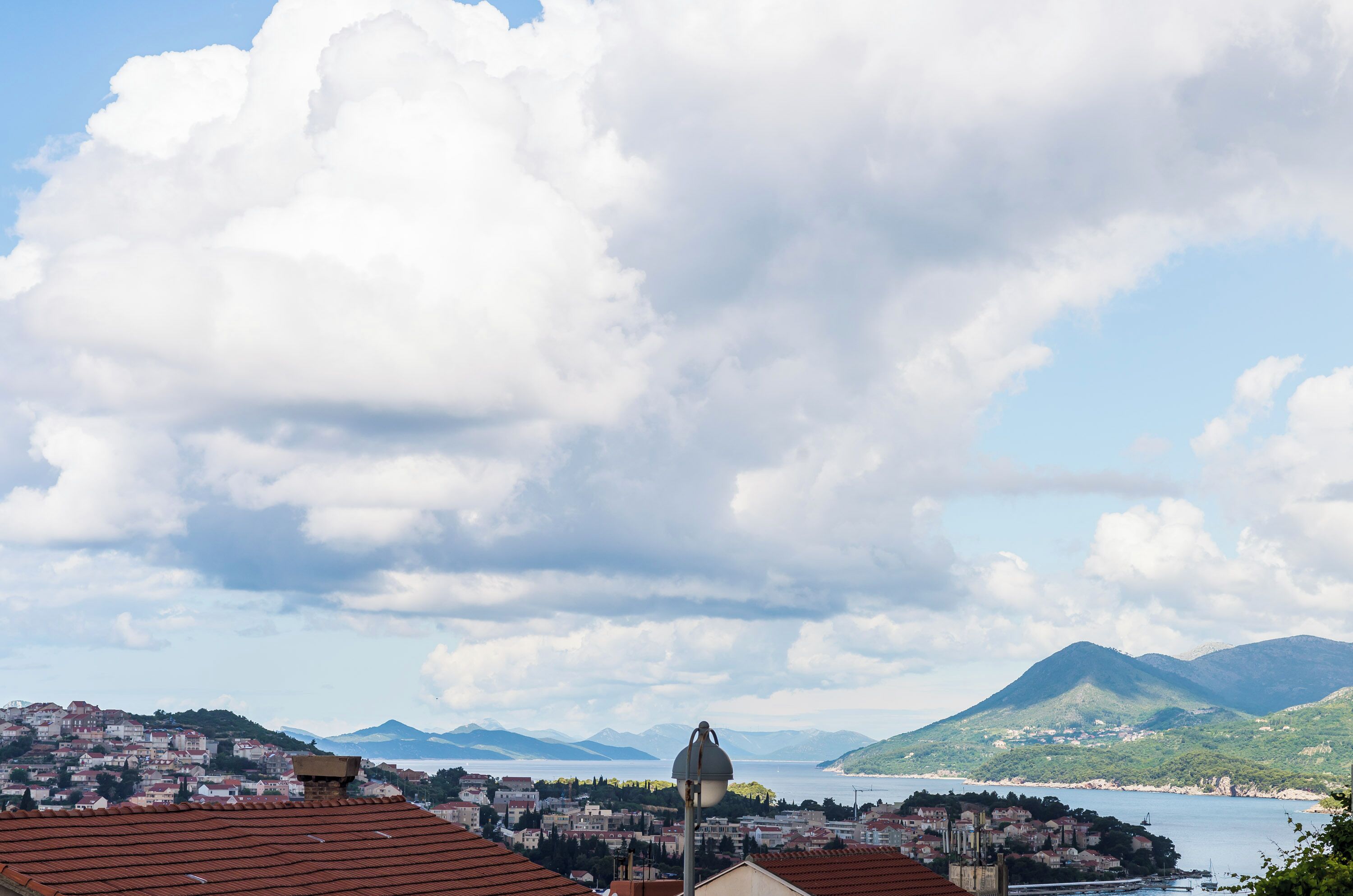 Uma vila costeira com edifícios de telhados vermelhos, céu claro e montanhas ao fundo.
