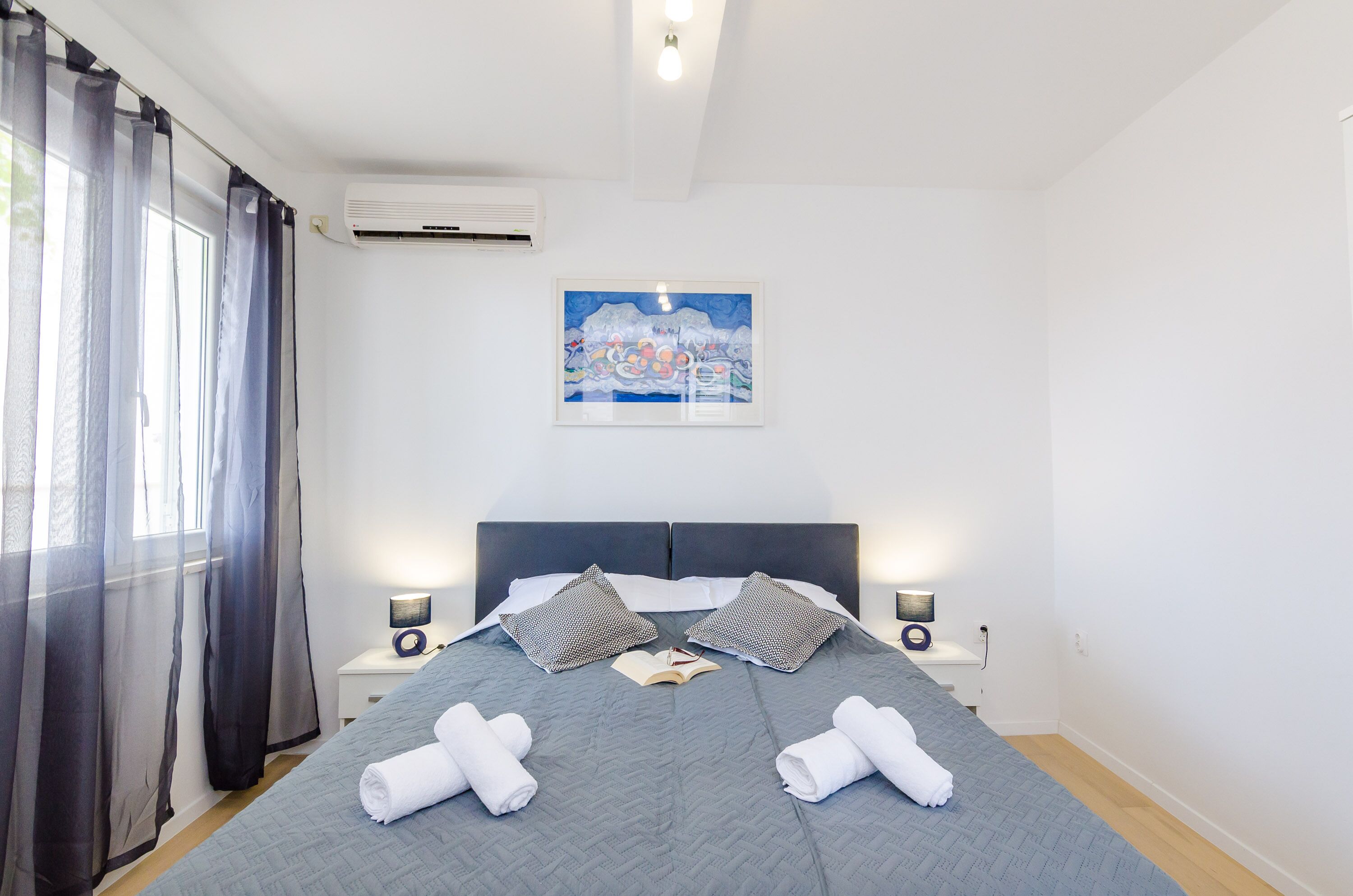 Een netjes opgemaakt bed met kussens en handdoeken, een nachtkastje met een lamp, en een schilderij aan de muur.