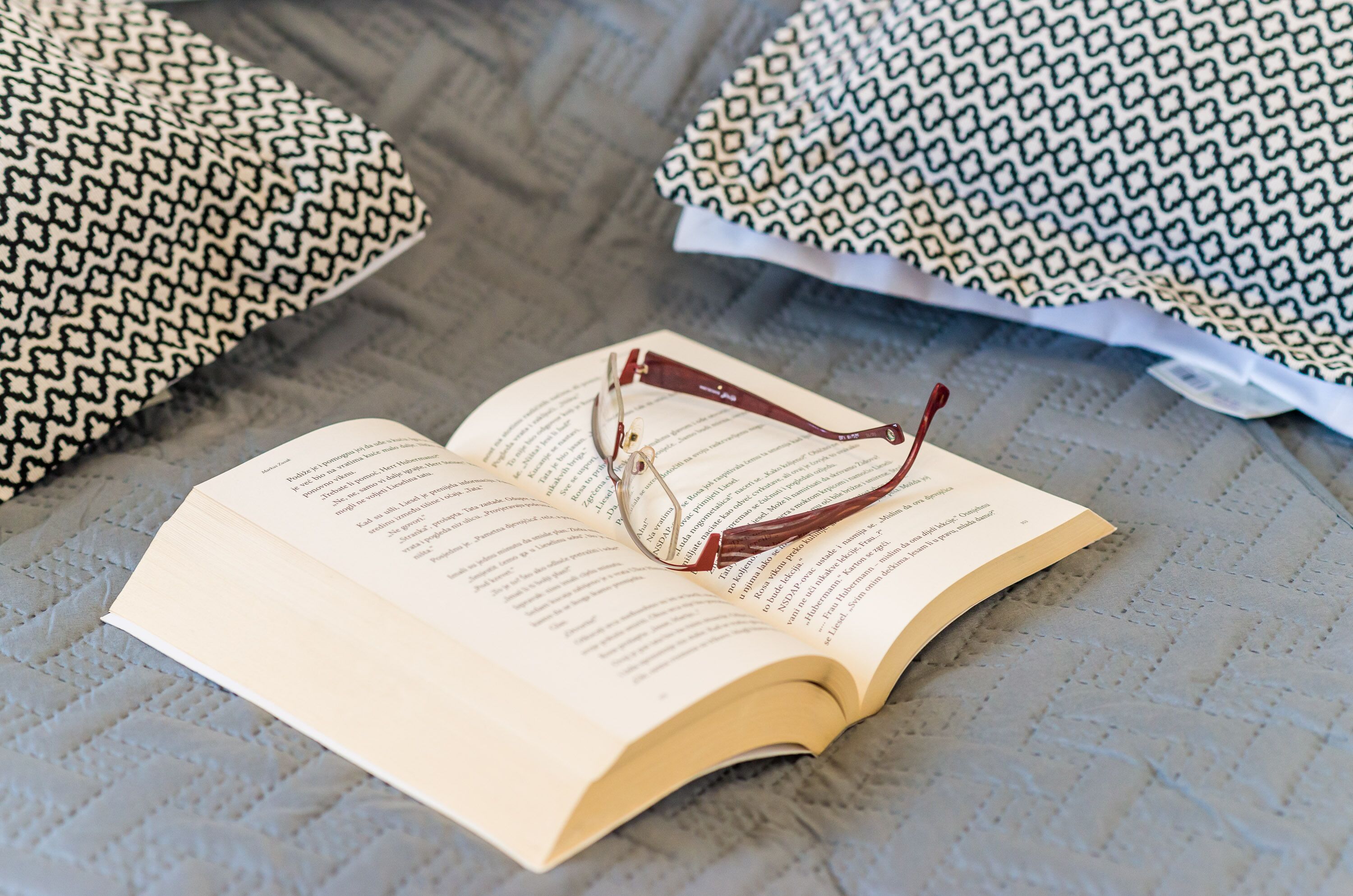 An open book with glasses on top, placed on a textured surface with patterned pillows in the background.