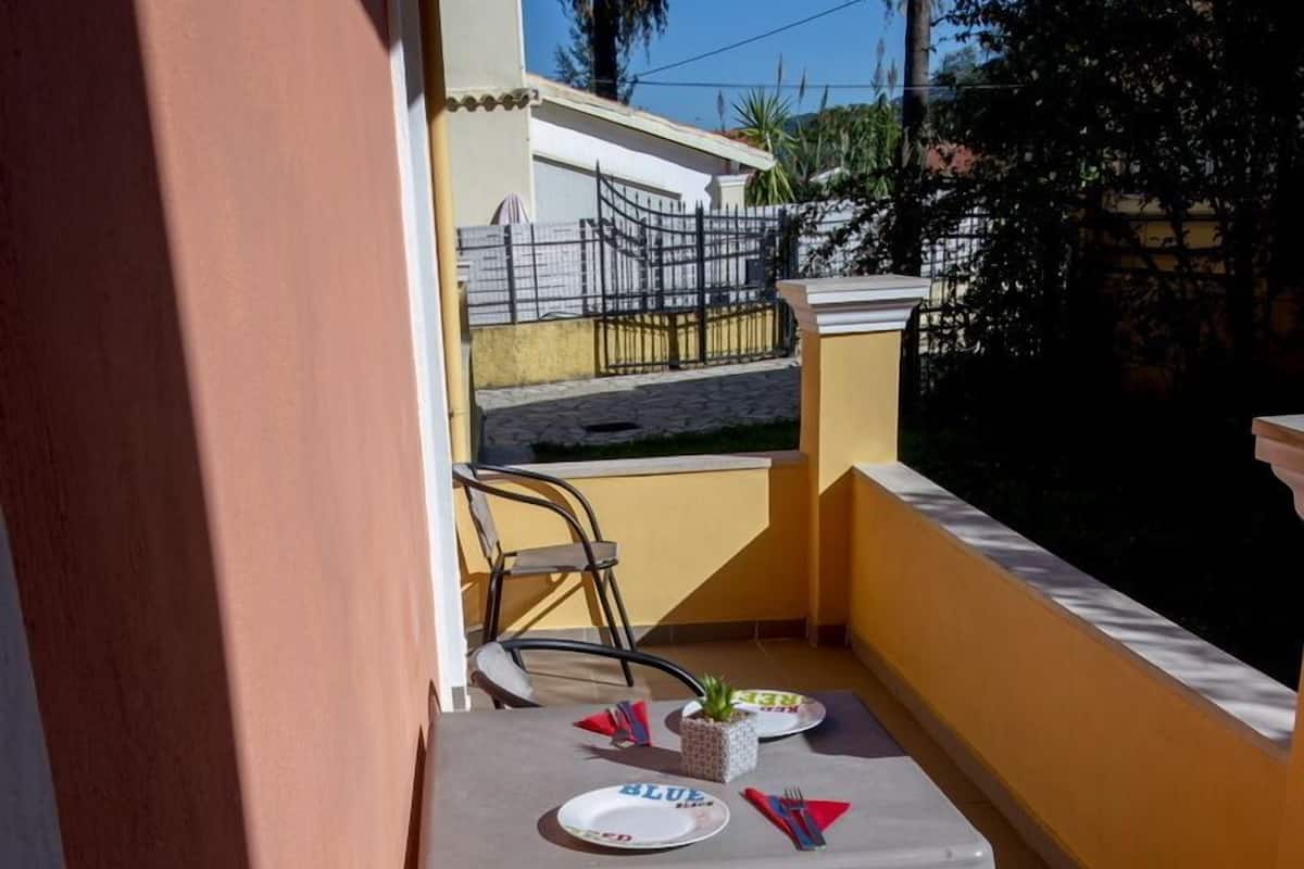 A small balcony with a table and chairs, a potted plant, and a view of a neighboring building and palm trees.