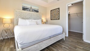 A bedroom with a large bed, two bedside lamps, a decorative headboard, a painting above the bed, and a walk-in closet with a metal shelf. - Spacious Coastal Home w/ Close Beach Access & Private Pool in Destin! 🌊🌴☀ (Destin)