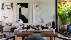 Een moderne woonkamer met een bank, salontafel en stoelen. Drie grote schilderijen hangen aan de muur en er staat een potplant in de hoek.
