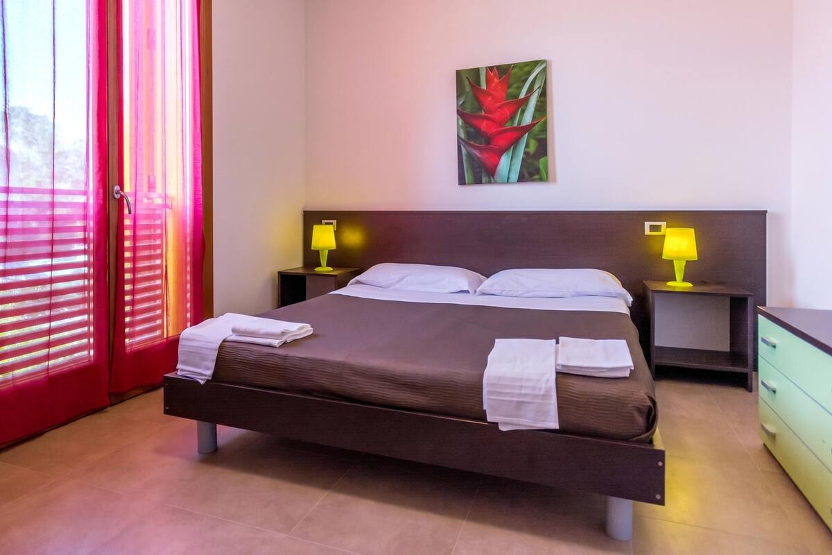 Un lit double avec du linge de lit blanc, deux tables de chevet avec des lampes, un tableau au mur et une porte rouge avec des stores.