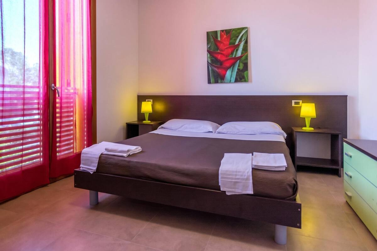 Un lit bien fait, avec du linge de lit blanc, deux tables de chevet avec des lampes, un tableau au mur et une porte coulissante avec des stores rouges.