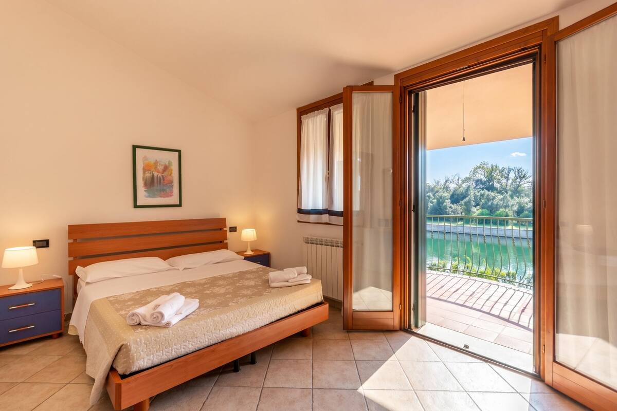 Une chambre à coucher avec un lit, des tables de chevet, une vue sur un balcon doté d’une balustrade et une fenêtre avec des rideaux.