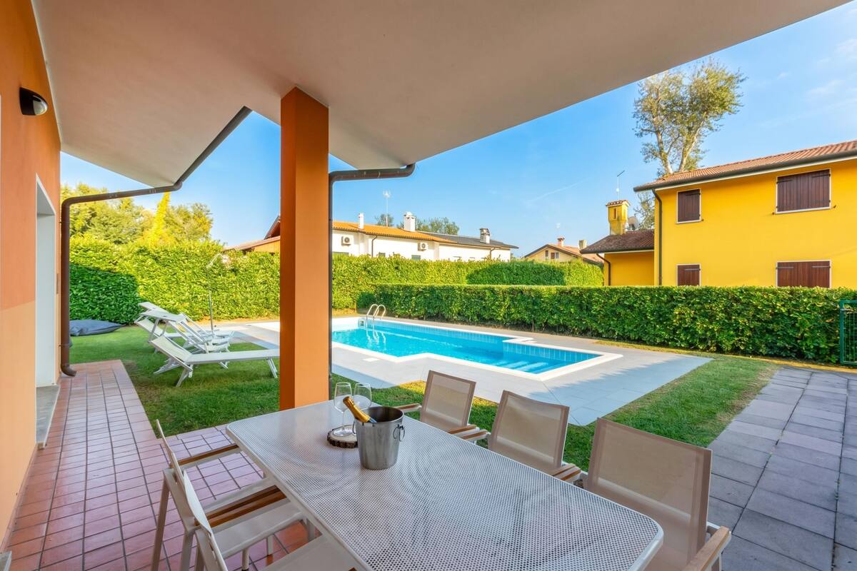 Une terrasse avec une table, des chaises et une bouteille de champagne. Au fond, on aperçoit une piscine et une haie.