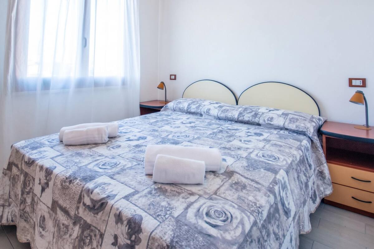 Un lit bien fait, recouvert d’une couvre-lit à motifs, avec deux serviettes enroulées, une table de chevet avec une lampe et une fenêtre ornée de rideaux légers.