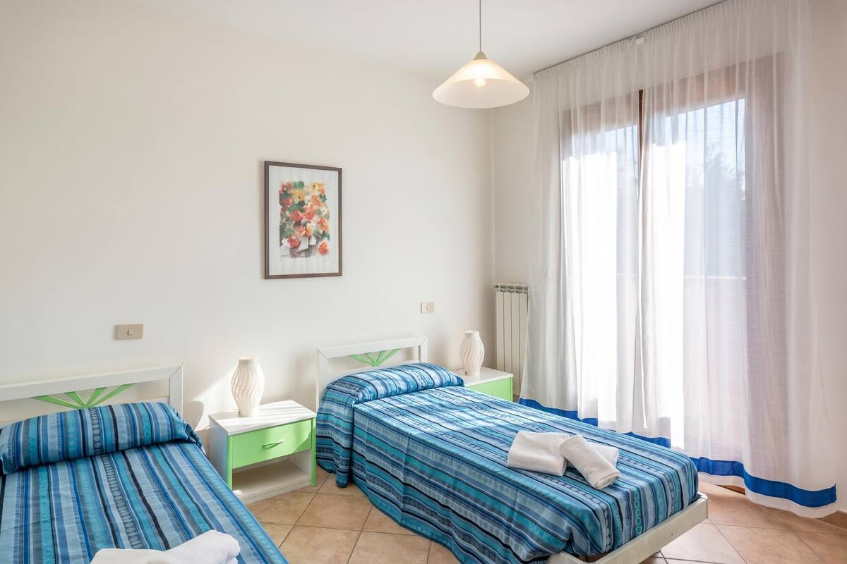 Une chambre d’hôtel avec deux lits, une table de chevet avec une lampe, un tableau encadré au mur et une grande fenêtre avec des rideaux légers.