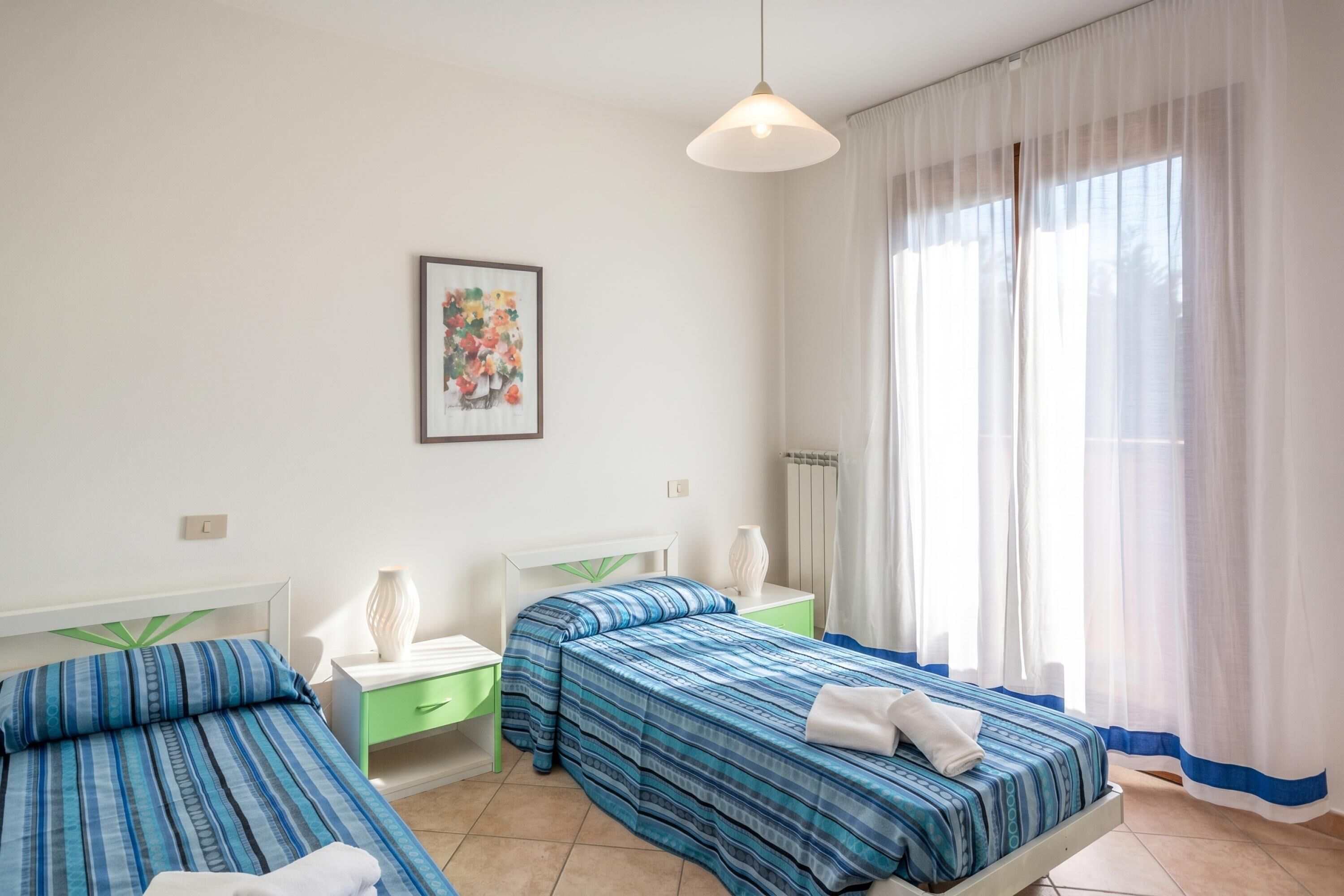 Ein Hotelzimmer mit zwei Betten, einem Nachttisch mit Lampe, einem Fenster mit durchscheinenden Vorhängen und einem gerahmten Bild an der Wand.