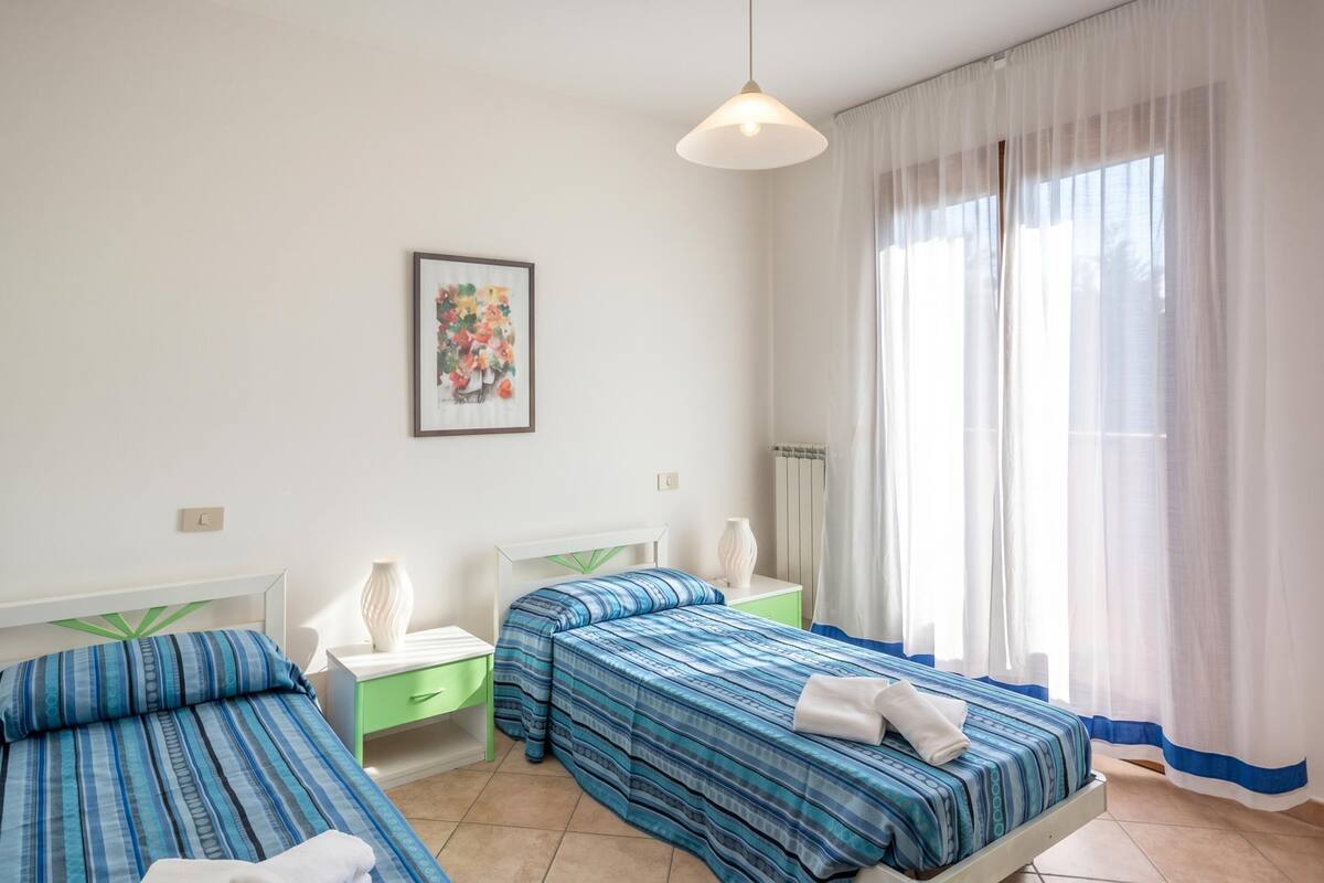 Une chambre d’hôtel avec deux lits, une table de chevet, une lampe, un tableau encadré et une fenêtre avec des rideaux légers.