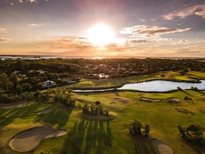 An aerial view of a golf course with a lake, trees, and houses in the background. - Albarella (Rosolina)