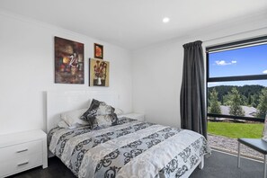 A bedroom with a large bed, a nightstand, a dresser, and a window with a view of a green landscape. - Mapua View - Mapua Holiday Unit (Mapua)
