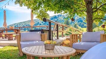 An outdoor seating area with a round wooden table, two cushioned chairs, a wicker basket with fruit, and a view of a forested hillside.