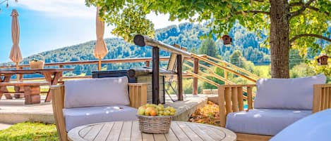 An outdoor seating area with a round wooden table, two cushioned chairs, a wicker basket with fruit, and a view of a forested hillside.