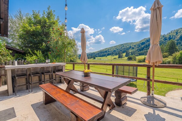 An outdoor dining area with a wooden table and benches, surrounded by a wooden fence, overlooking a green landscape with hills.