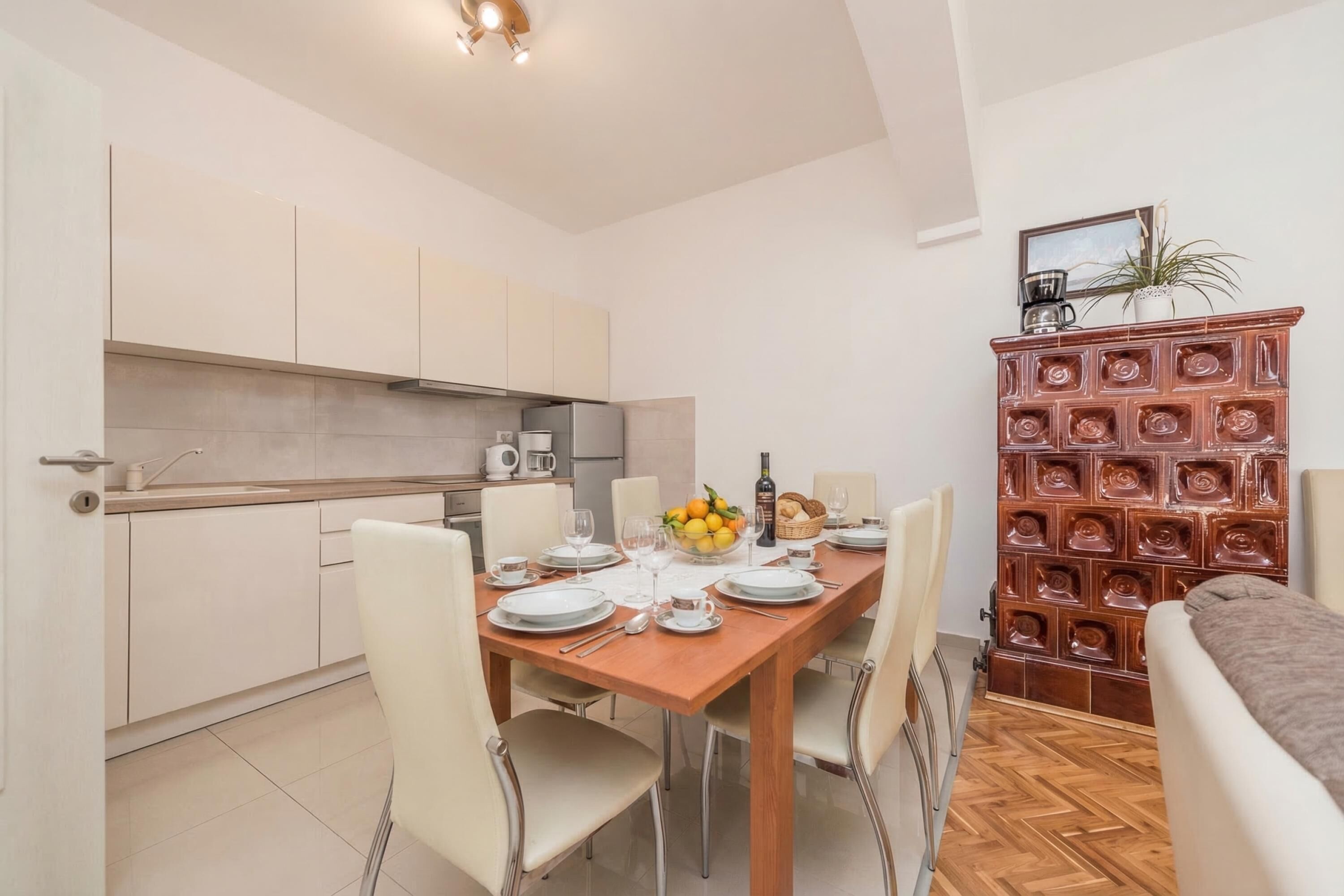 Een eetruimte met een houten tafel voor vier personen, witte stoelen, een keuken op de achtergrond en een decoratieve houten kast met een plant erop.