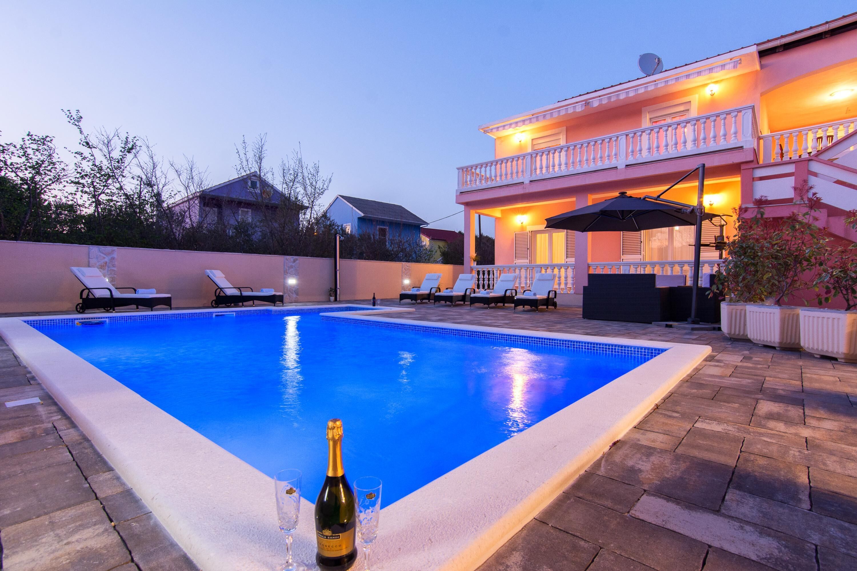 En swimmingpool med liggestole, en champagneflaske og et toetagers hus med balkon.