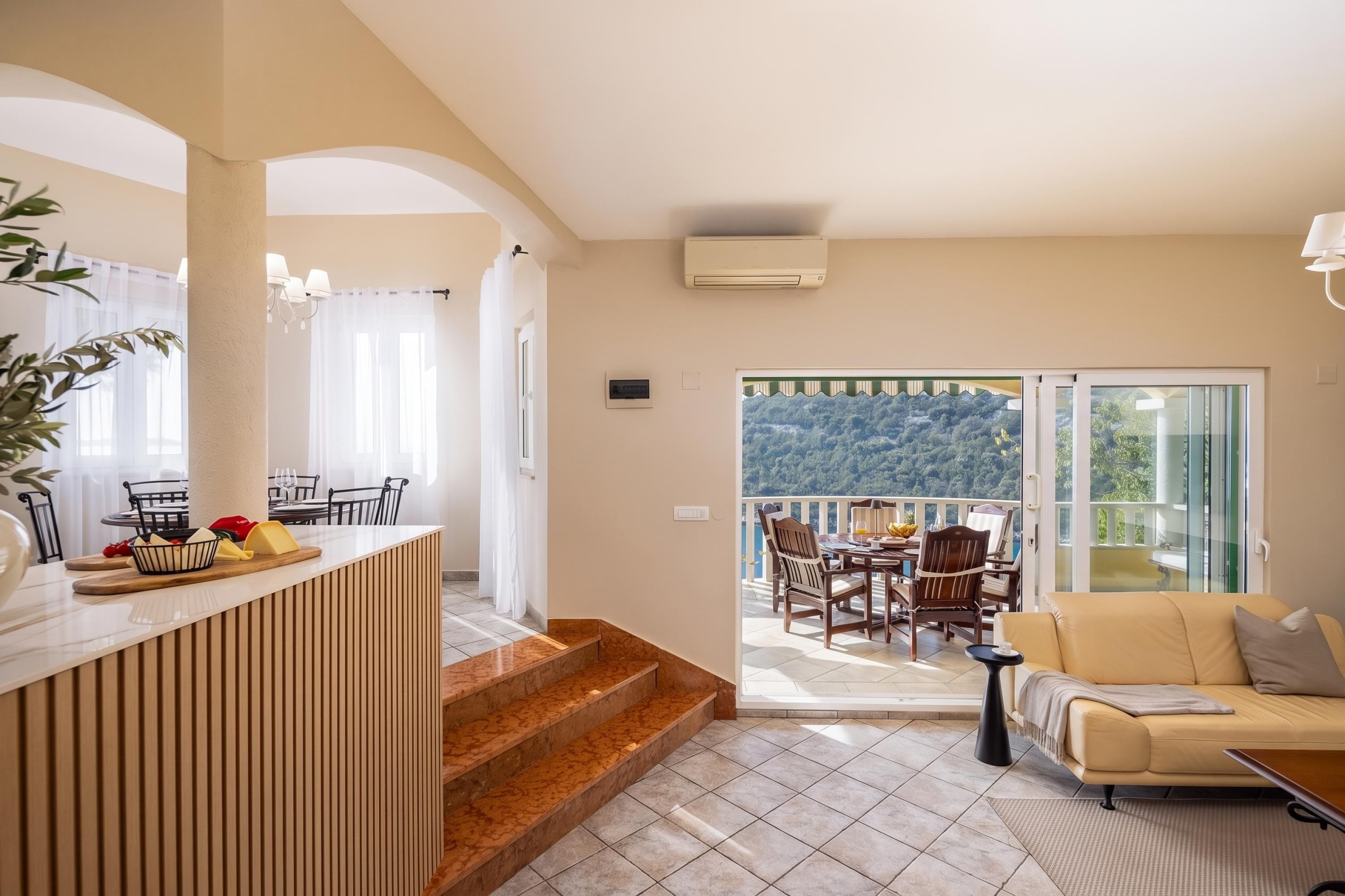 A modern living room with a sofa, a dining area with chairs and a table, a kitchen area with a counter and a plant, and a balcony with a view of the mountains.