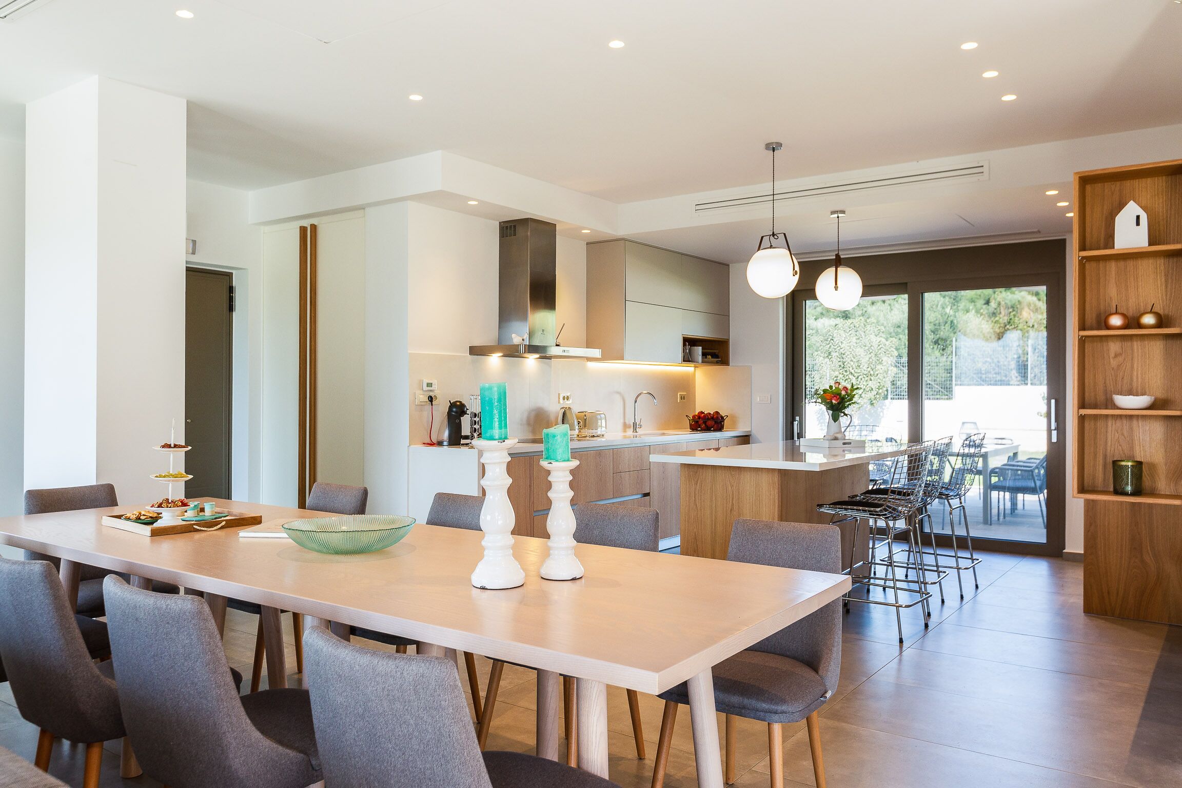A modern kitchen with a central island, a dining area with a long table and chairs, and a view of the outdoors through large windows.