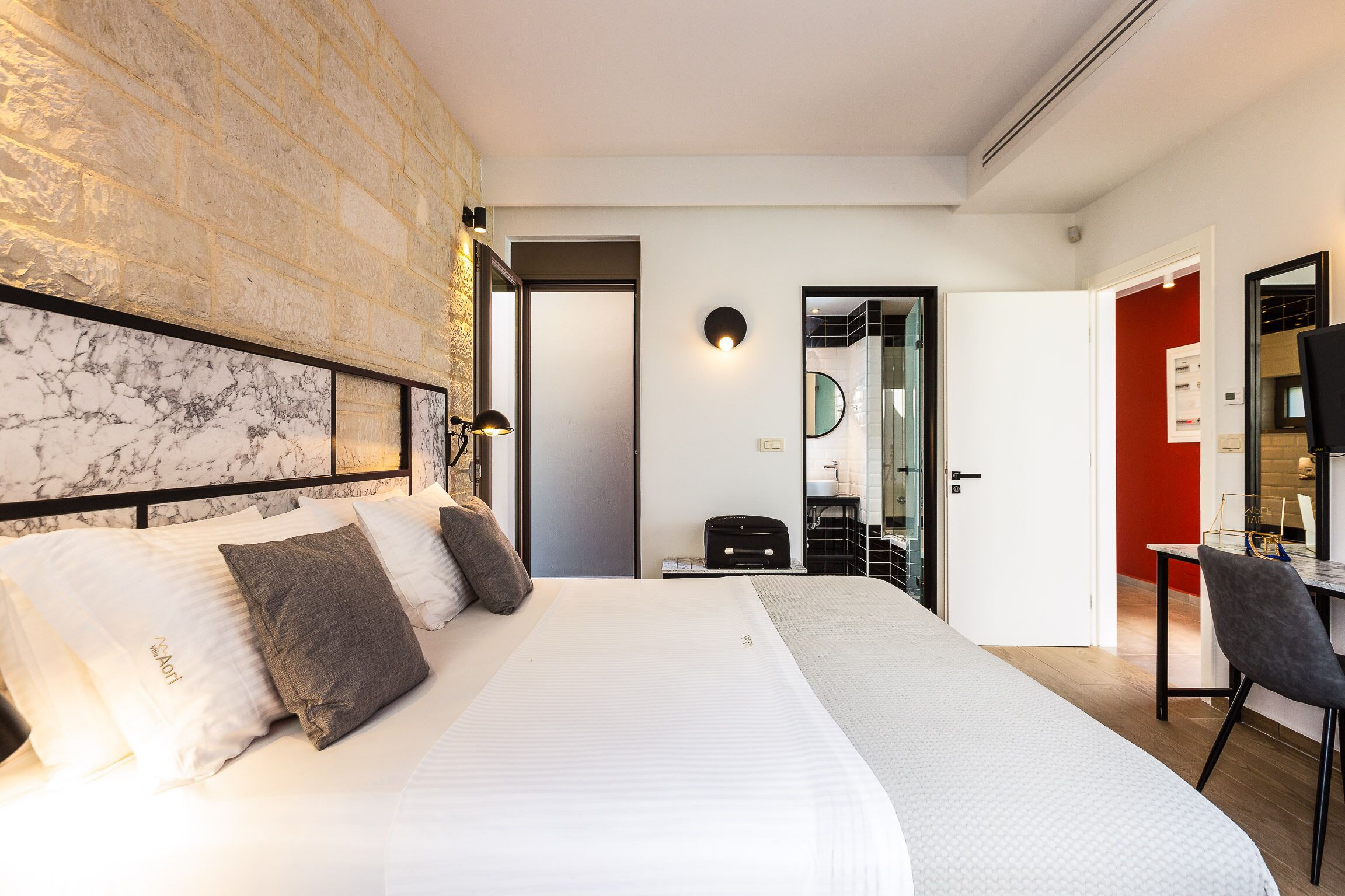 Habitación de hotel con una cama grande, cabecera de mármol, un escritorio con silla, y un baño visible a través de una puerta abierta.
