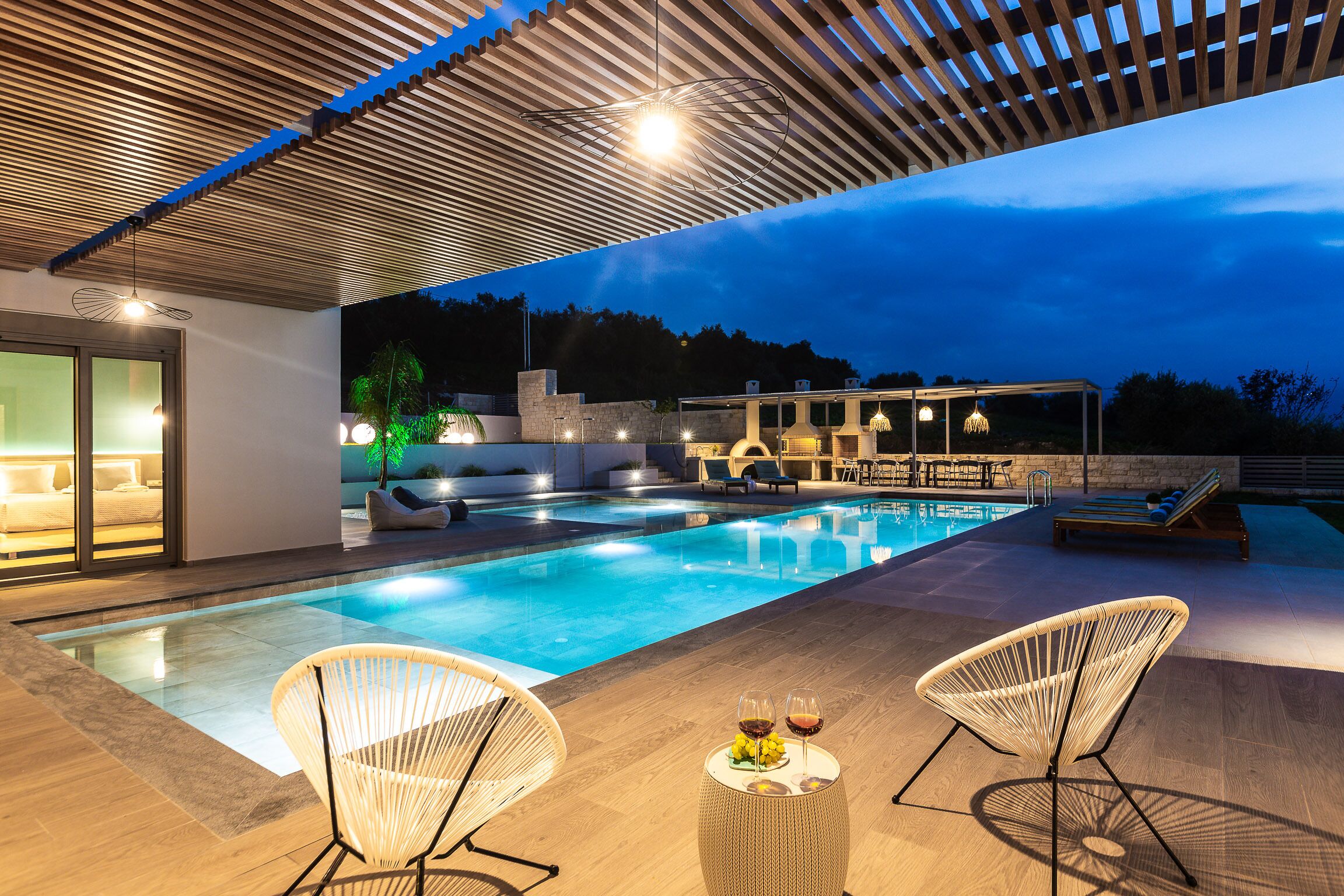Area piscina moderna all'aperto con due lettini, un tavolo con bevande e una zona relax coperta illuminata.