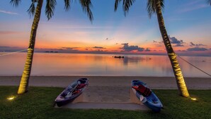 Escena de playa con dos kayaks y una caña de pescar, palmeras y una puesta de sol tranquila sobre el agua.