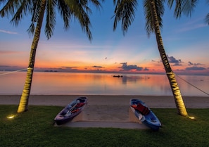 A beach scene with two kayaks and a fishing rod, palm trees, and a calm sunset over the water. - Baan Asan (Koh Samui)