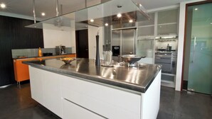 A modern kitchen with a large island, stainless steel appliances, and a glass enclosure over the stove. - Baan Asan (Koh Samui)