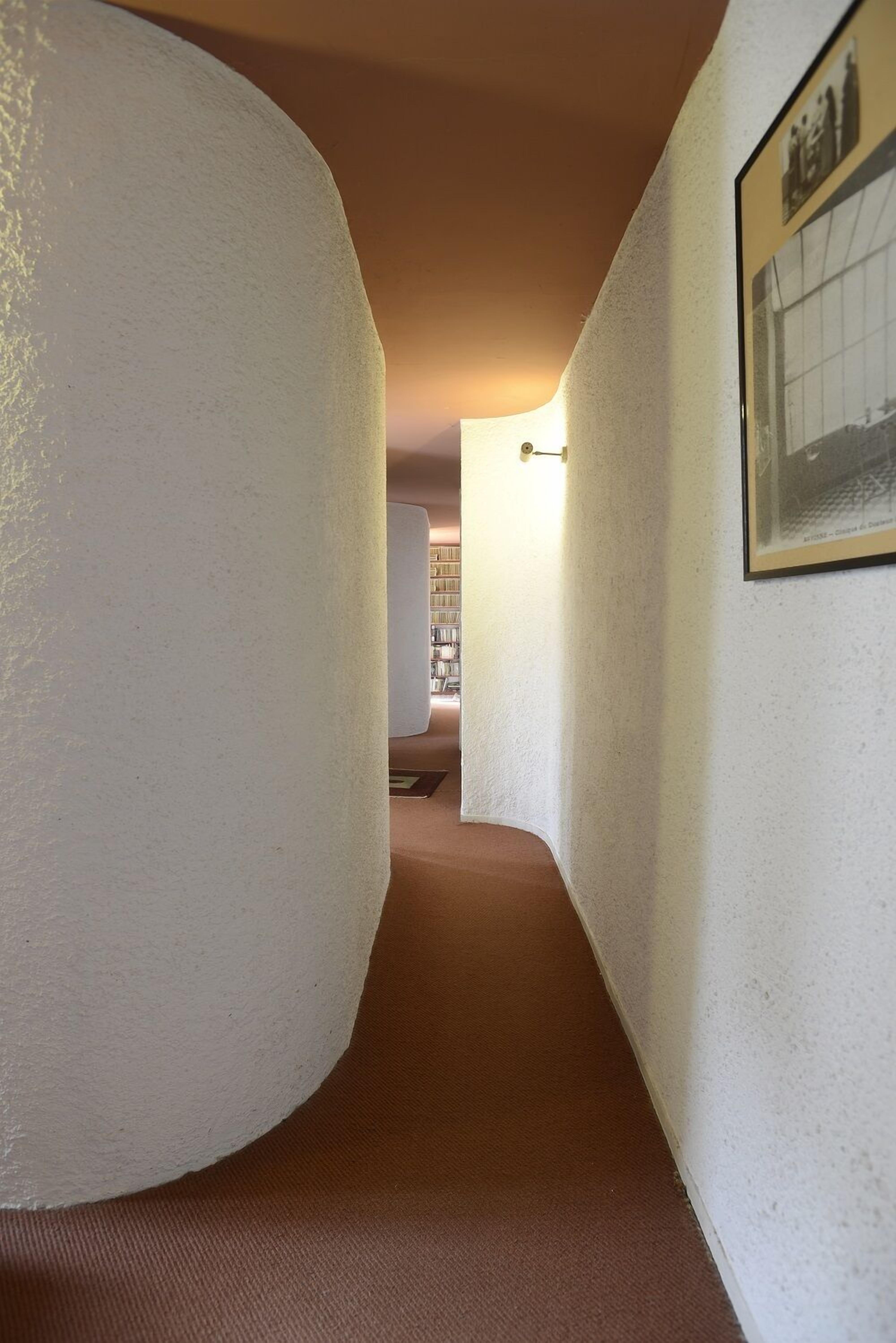 Ein Flur mit weißen Wänden und Teppichboden, der zu einem Raum mit Bücherregal und einem gerahmten Bild an der Wand führt.