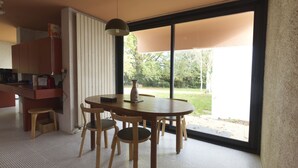 Un comedor con una mesa redonda y cuatro sillas, un florero sobre la mesa, y vistas a un jardín a través de grandes ventanales.