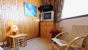 A cozy room with a wooden wall, a TV on a cabinet, a small table with a lamp, and a woven chair. - Cozy studio at the slopes with balcony, pets welcome (Chamrousse)