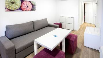 Una sala moderna con un sofá gris, una mesa blanca y un otoman rojo. Hay una obra de arte colorida en la pared y un aparador blanco en la esquina.