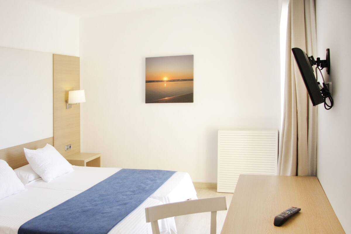 A hotel room with a bed, a bedside table, a desk, a chair, a lamp, a picture of a sunset, a phone, and a window with curtains.