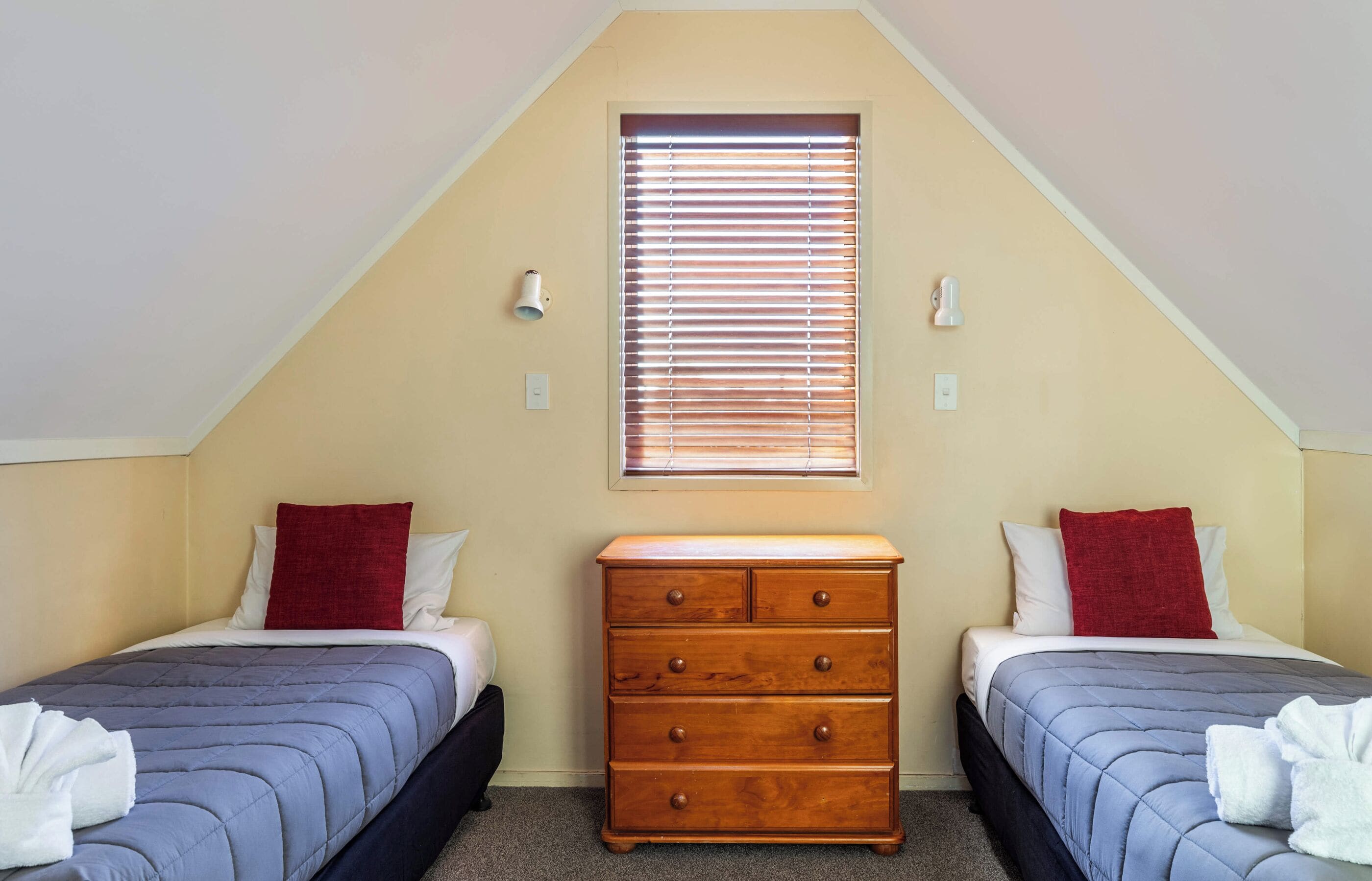 Une chambre avec deux lits, une commode, une fenêtre avec des stores et un luminaire.