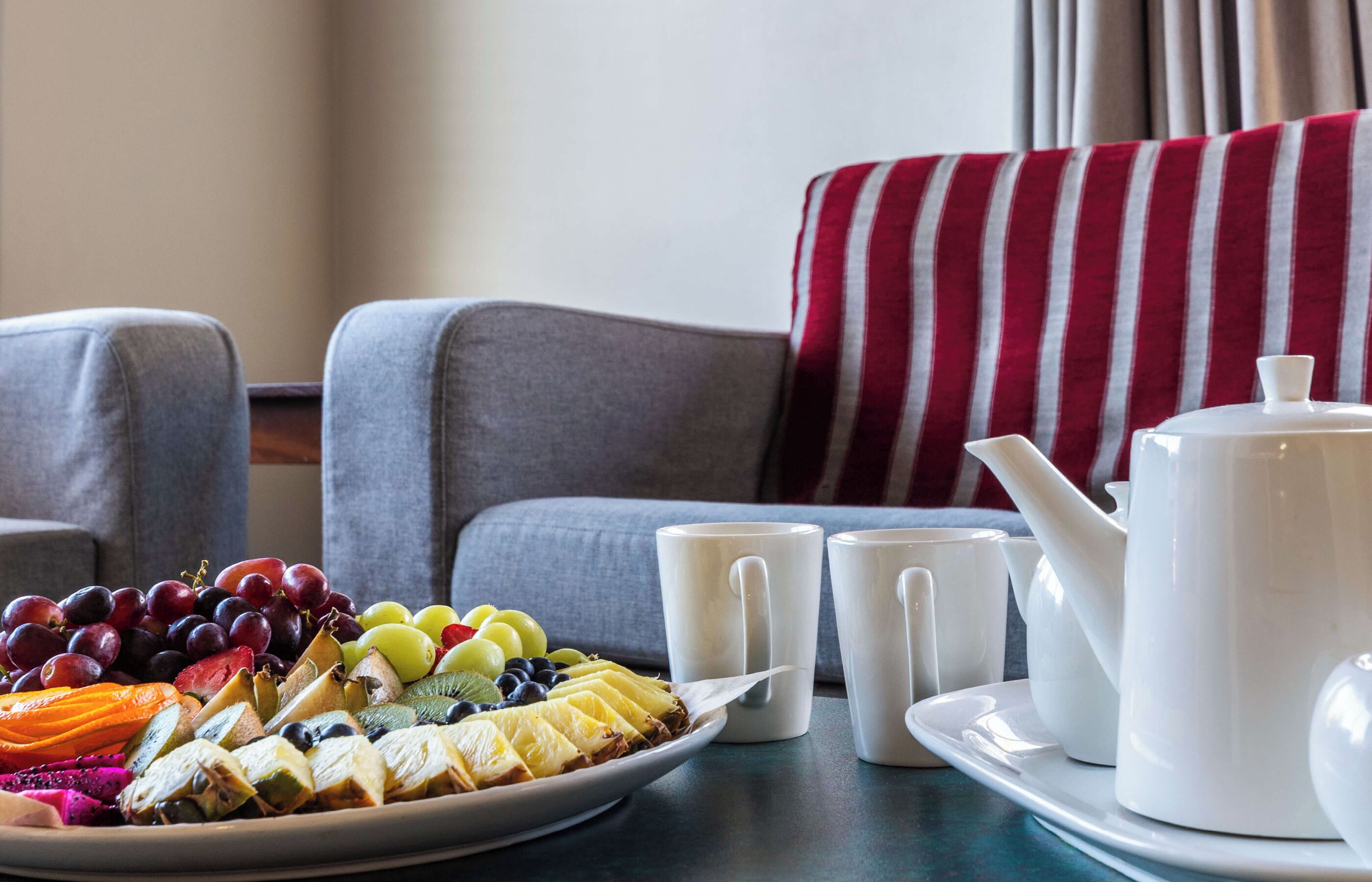 A breakfast setting with a fruit platter, white mugs, and a teapot on a table.
