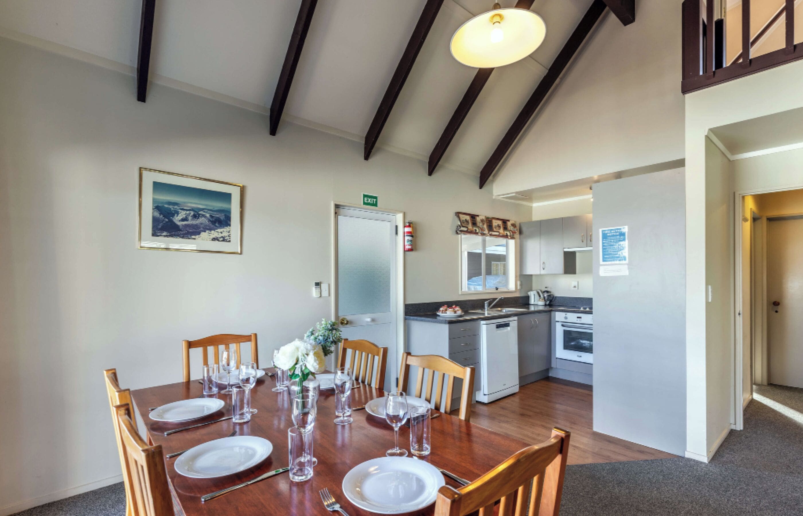 A dining area with a wooden table set for six, a kitchen in the background, and a framed picture on the wall.