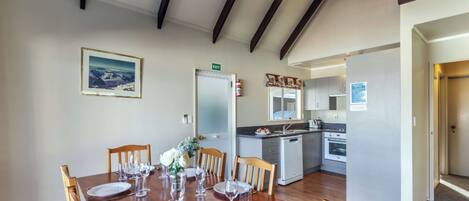 A dining area with a wooden table set for six, a kitchen in the background, and a framed picture on the wall.