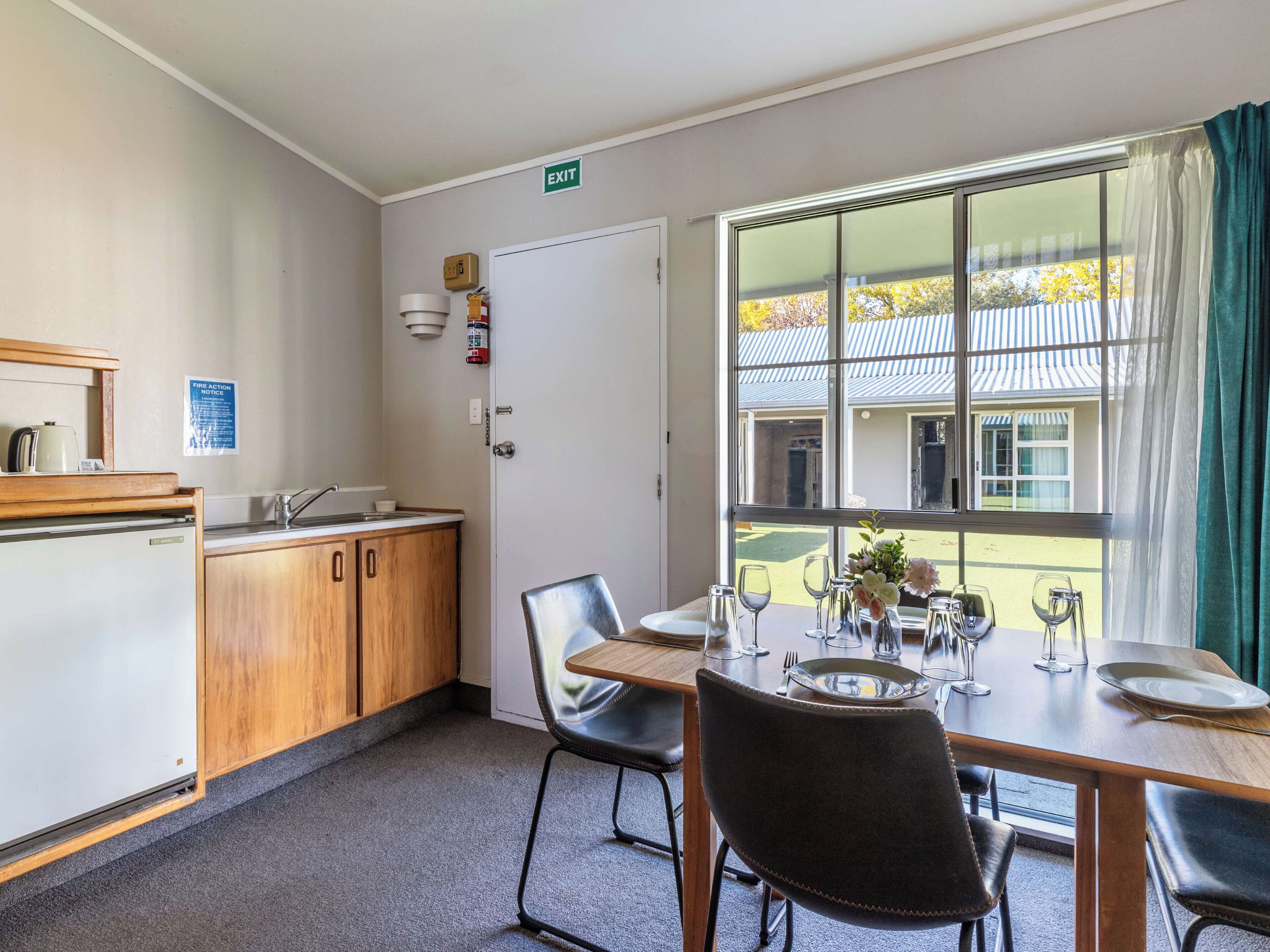Ein Essbereich mit einem Tisch für vier Personen, einem Kühlschrank, Holzschränken und einem Fenster mit Blick auf ein Gebäude und Bäume.