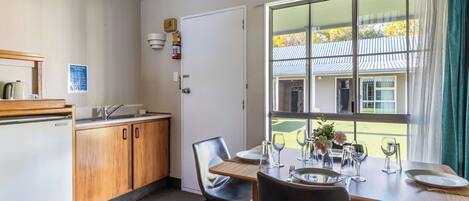 Ein Essbereich mit einem Tisch für vier Personen, einem Kühlschrank, Holzschränken und einem Fenster mit Blick auf ein Gebäude und Bäume.