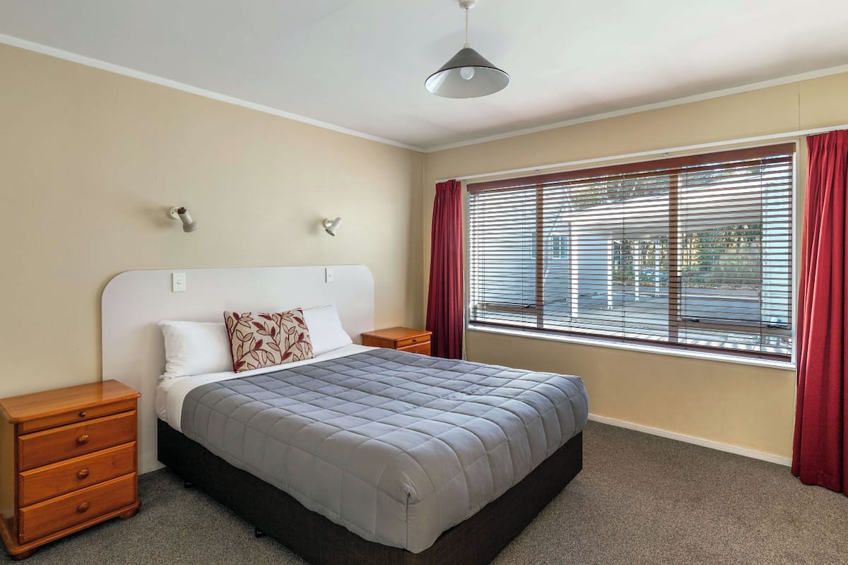 A bedroom with a bed, wooden nightstands, a ceiling light, a window with blinds, and a red curtain.