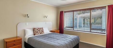 A bedroom with a bed, wooden nightstands, a ceiling light, a window with blinds, and a red curtain.