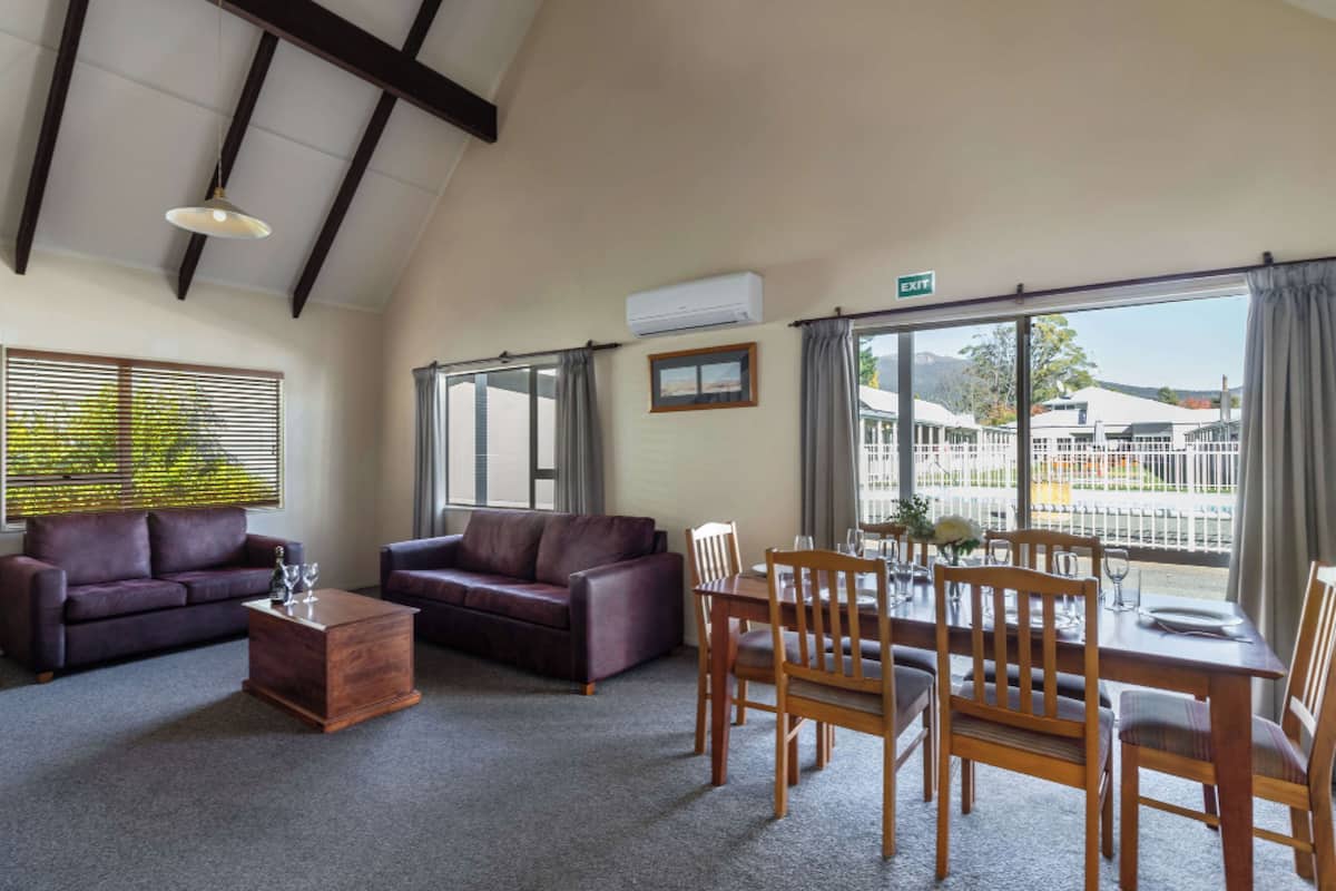 A spacious room with a high ceiling, a wooden dining table set for four, two leather sofas, a wooden coffee table, and a large window with curtains.