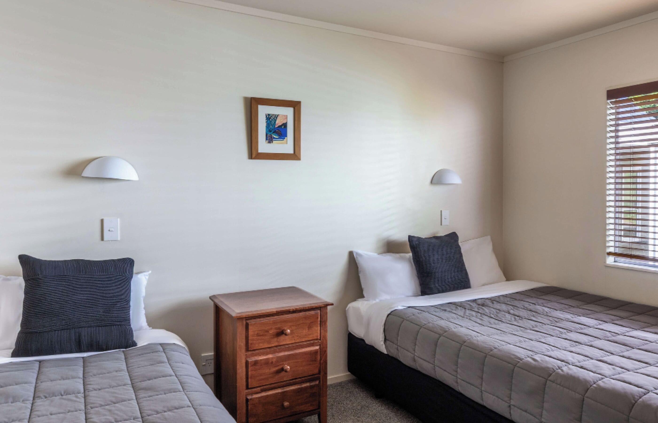 A bedroom with two beds, a nightstand, wall-mounted lamps, and a framed picture on the wall.