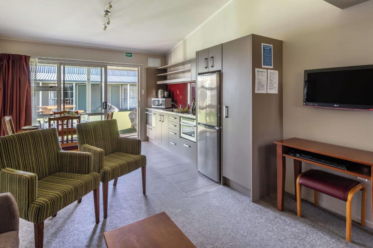 A hotel room with a kitchenette, a dining area with chairs, a TV mounted on the wall, and a window with curtains.