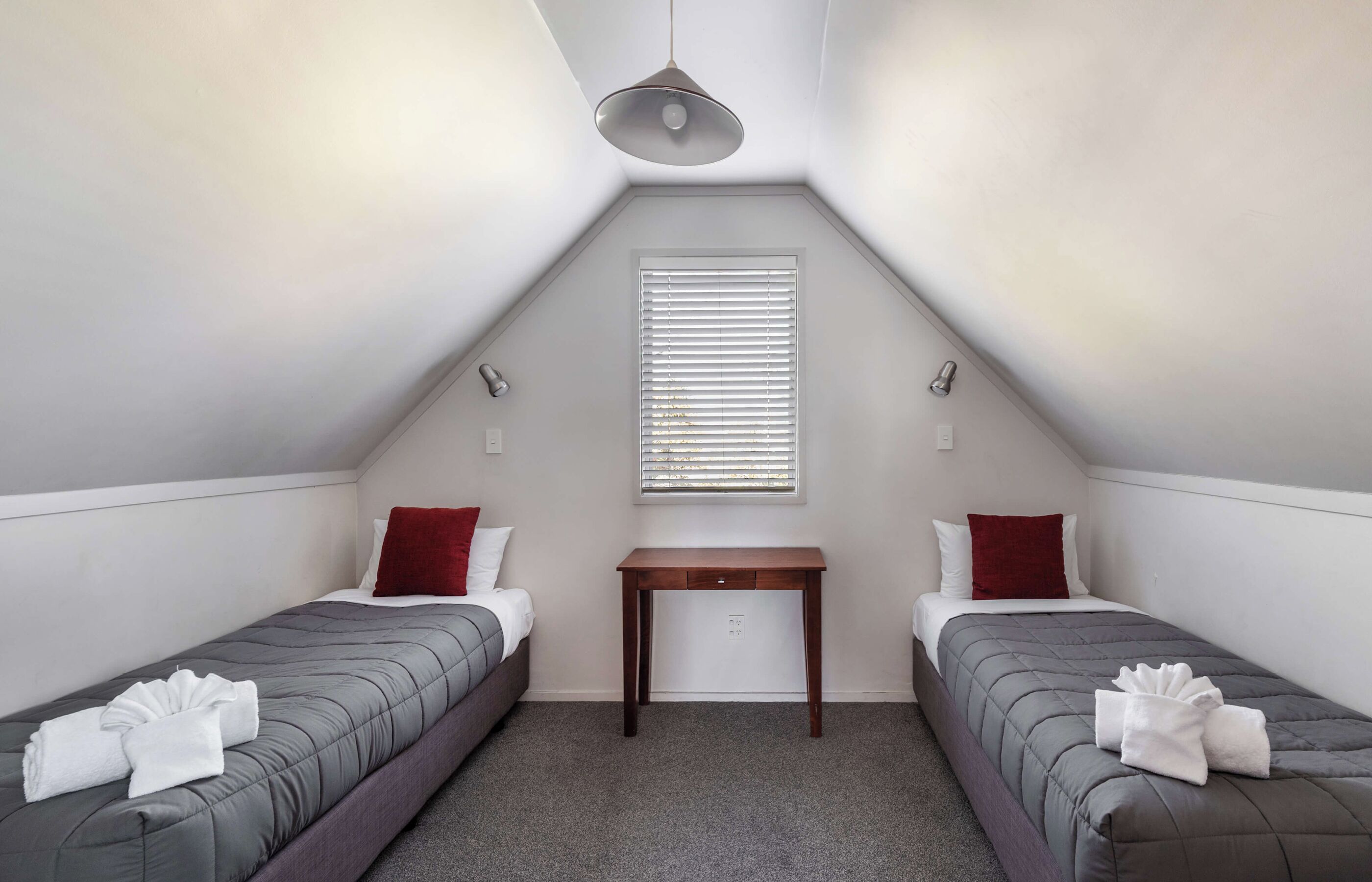 A small, triangular room with two single beds, a wooden table, and a window with blinds.