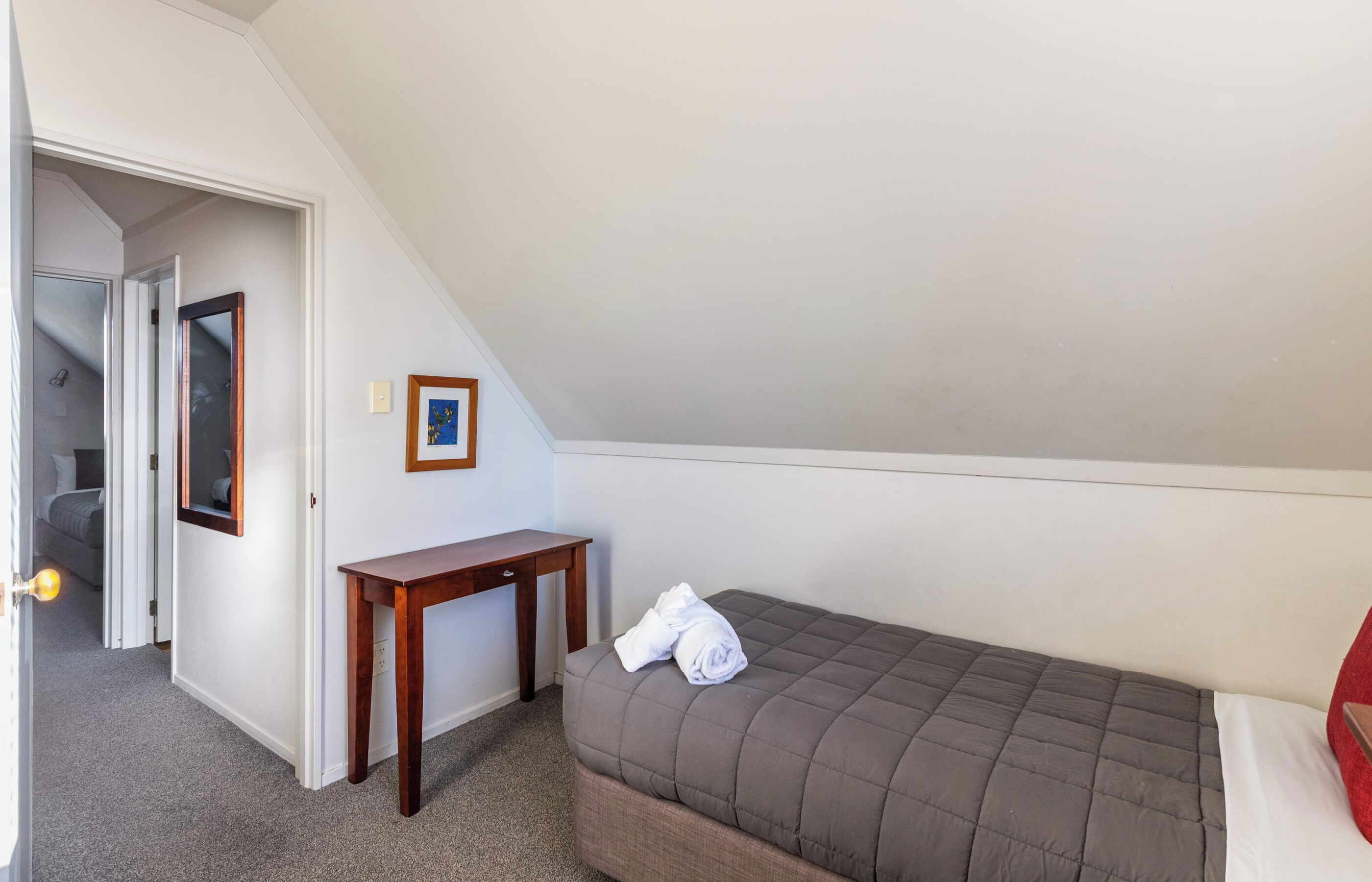 Quarto pequeno com uma cama de solteiro, teto inclinado, mesa de cabeceira de madeira, um quadro na parede e uma porta que dá para outro cômodo.