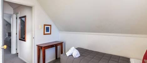 A small, single-bed room with a sloped ceiling, a wooden nightstand, a framed picture on the wall, and a door leading to another room.