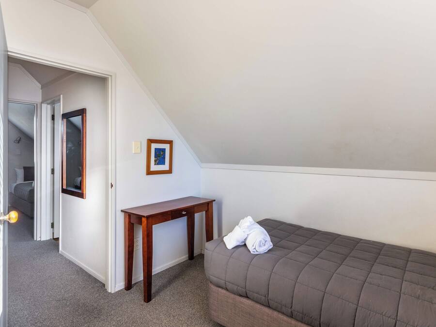 Quarto pequeno com uma cama de solteiro, teto inclinado, mesa de cabeceira de madeira, um quadro na parede e uma porta que dá para outro cômodo.