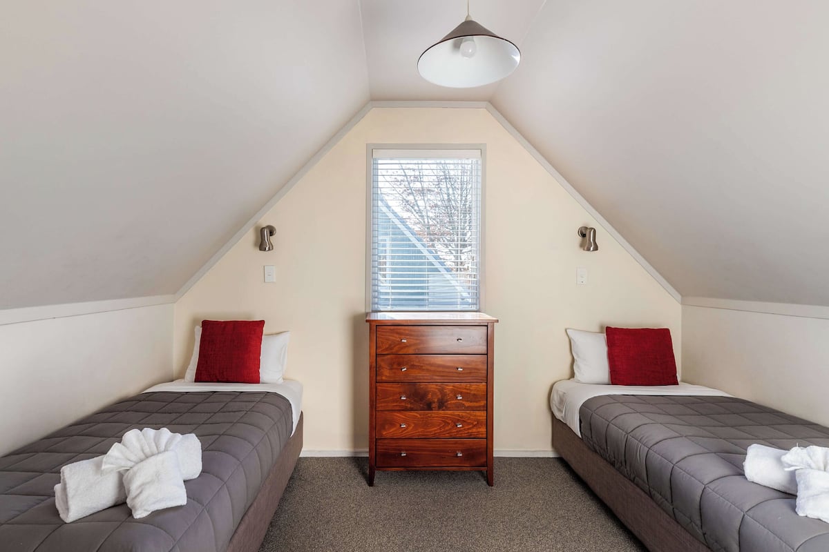 A small, triangular room with two beds, a wooden dresser, and a window with blinds.