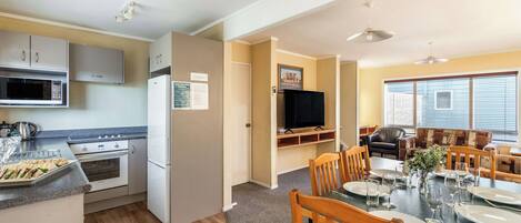 A compact kitchen with modern appliances, a dining area with wooden chairs, and a television in the living space.